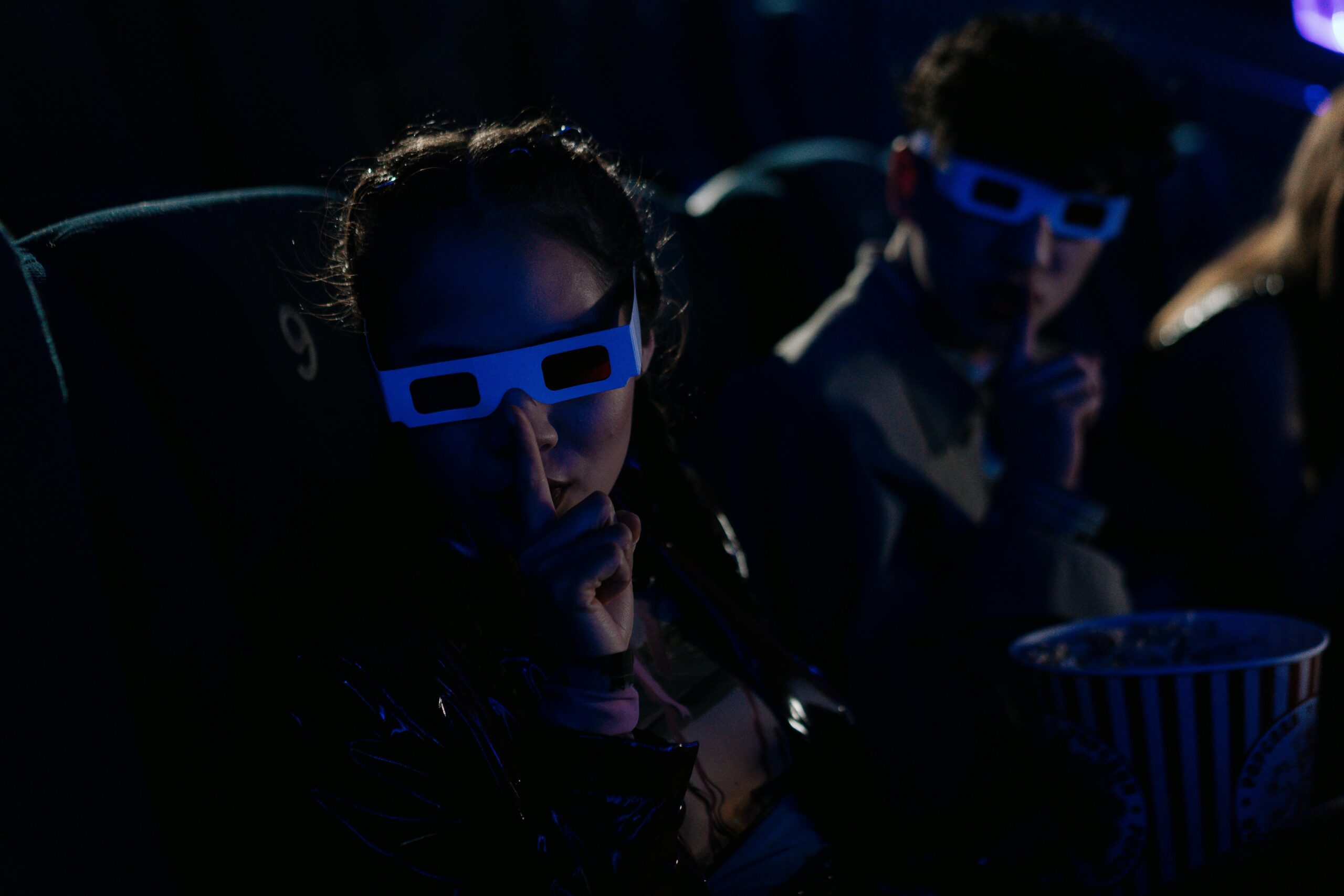 Teenagers wearing 3D glasses gesture to hush in a dimly lit cinema with popcorn.