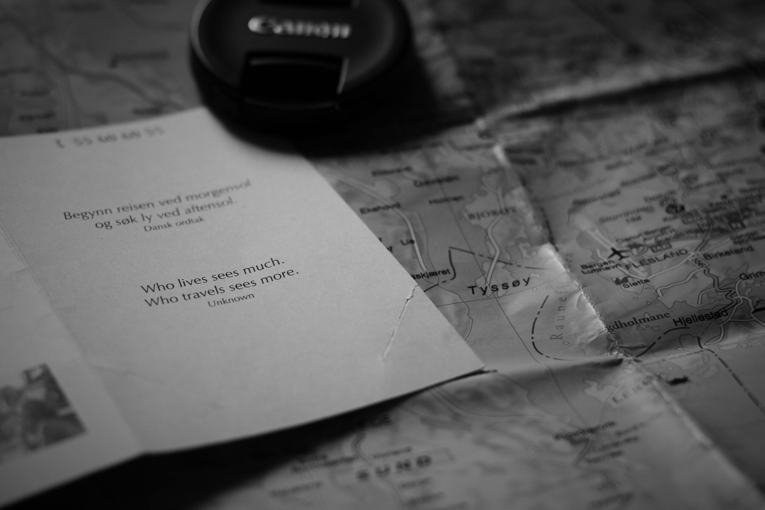 A black and white image of an inspirational travel quote alongside a map and camera lens cap.