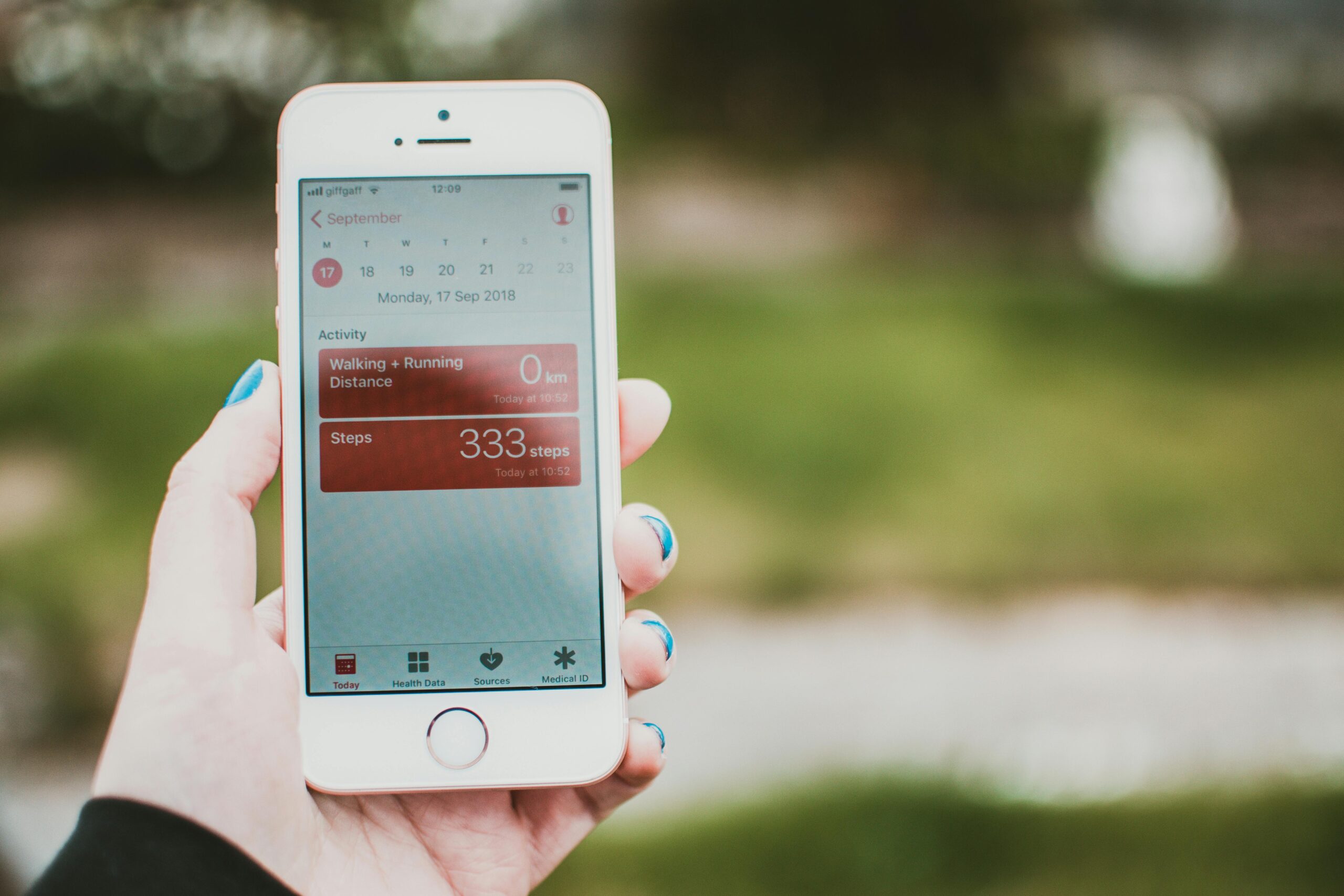 A person holds a smartphone showing a fitness app with step count and distance tracking outdoors.