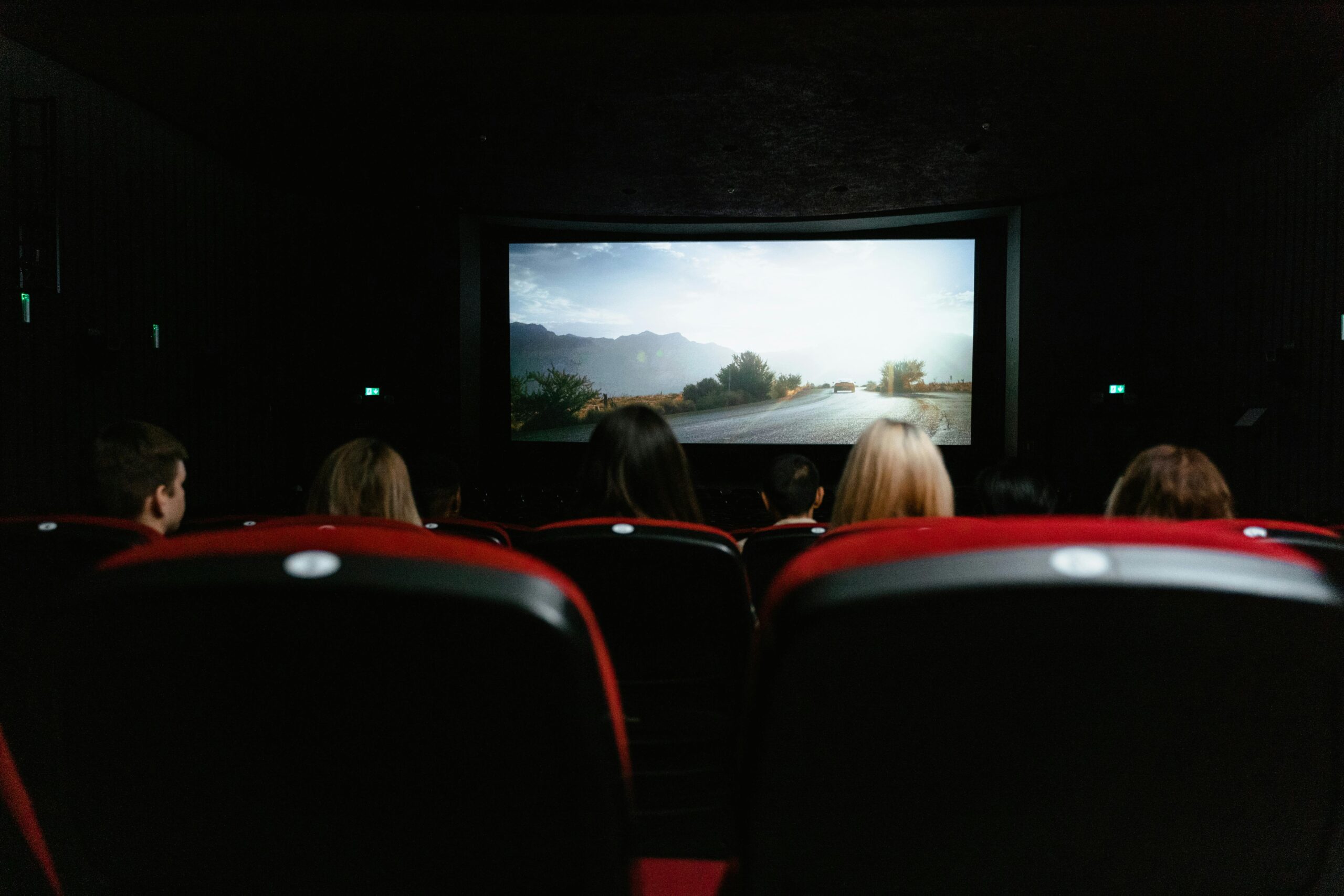 View from behind of people watching a movie in a cinema with red seats and a large screen.