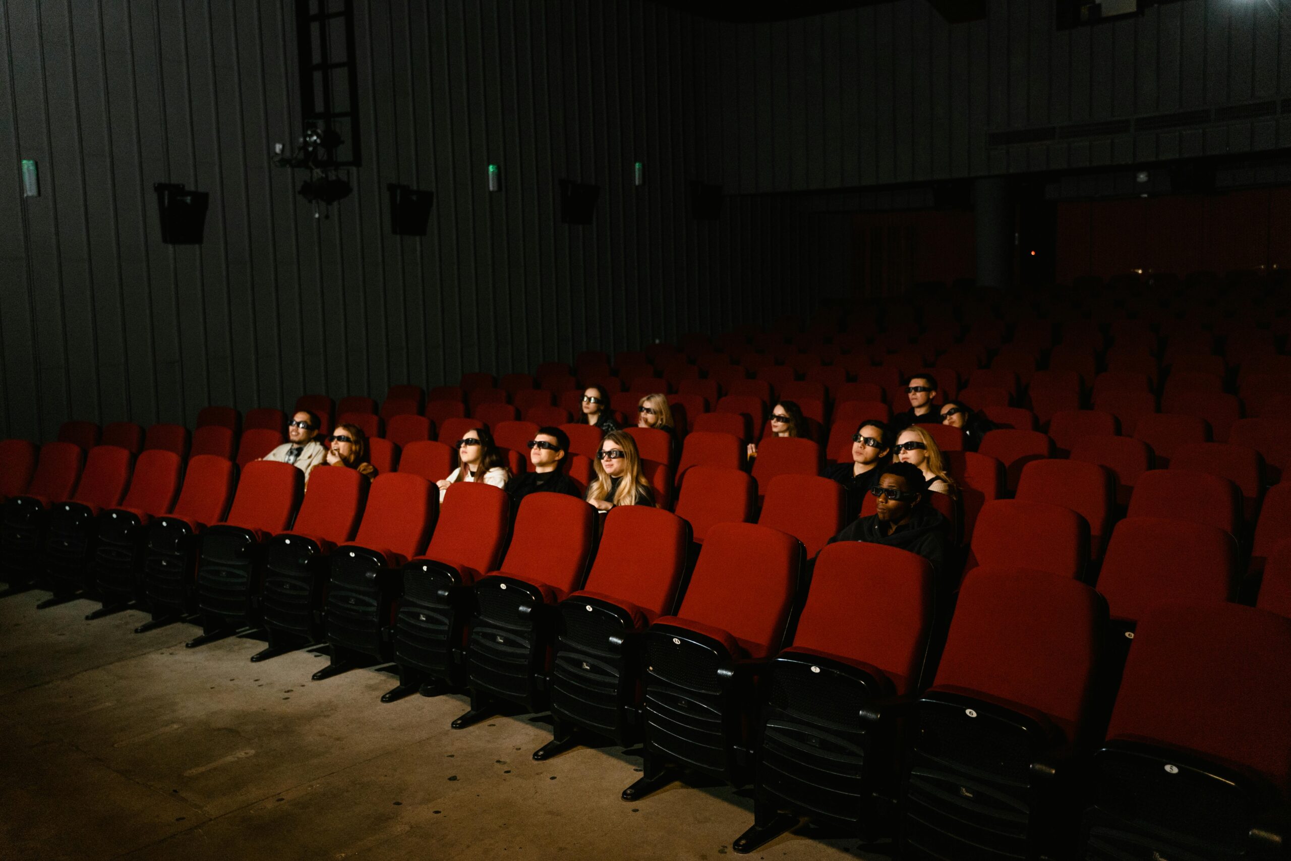Small group of people wearing 3D glasses watching a movie in a theater with red seats.