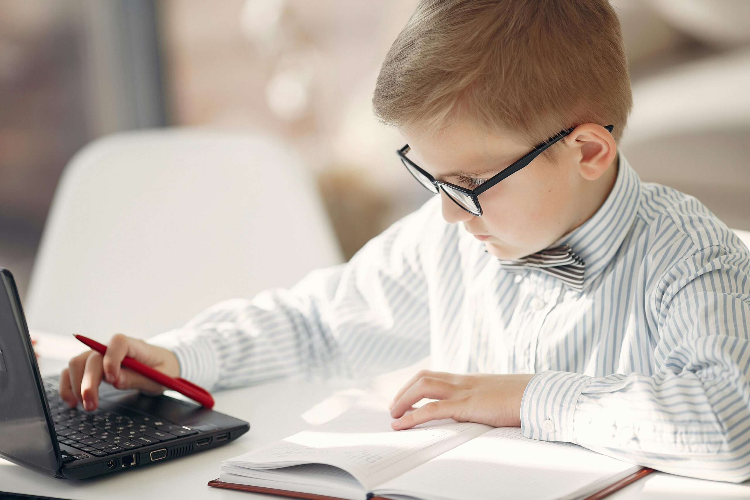 A young boy in glasses and bow tie diligently studies with a laptop at a table indoors.