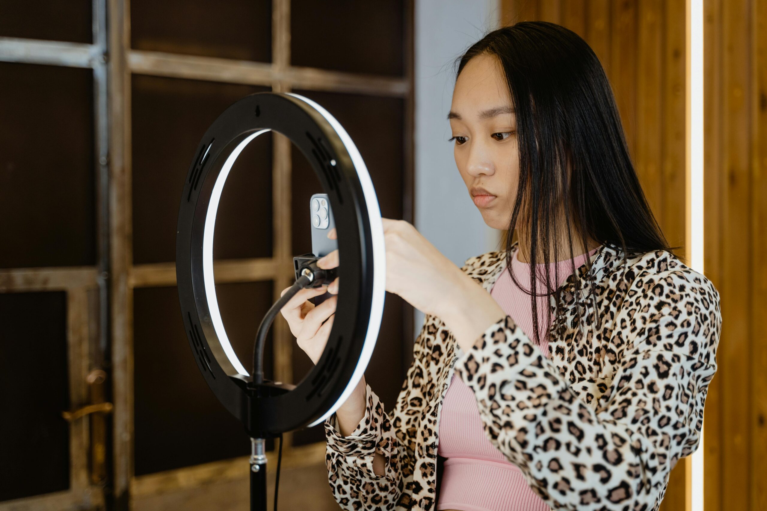 Asian woman with leopard print jacket adjusts ring light for smartphone streaming setup.